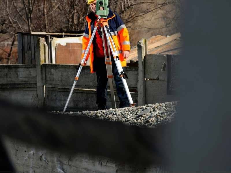 Construction worker in a bright safety jacket and helmet sets up a tripod-mounted surveying instrument at a gravel construction site.