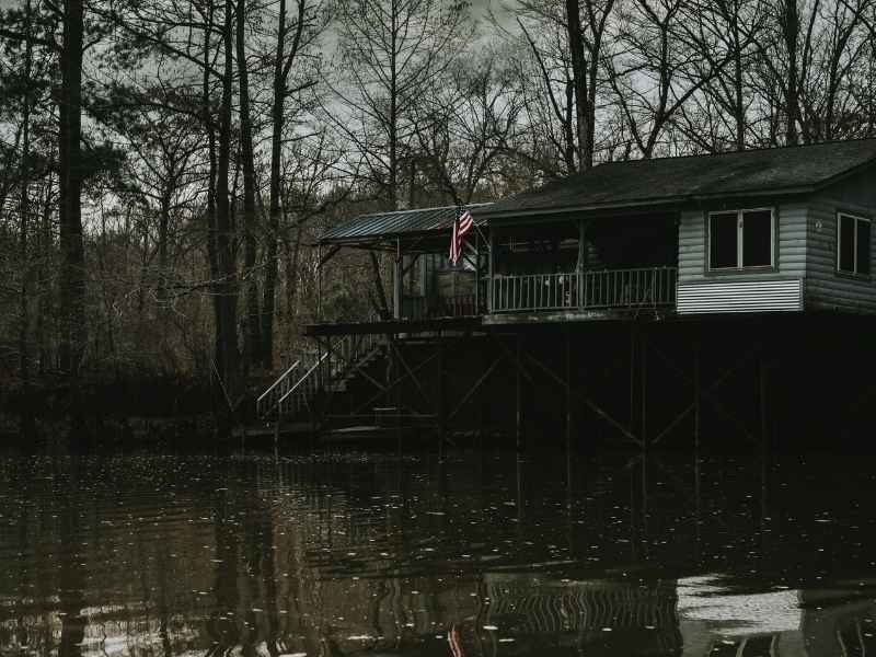 Wooden house on stilts over a dark lake, with a small porch and an American flag on the railing amid bare trees to the left.